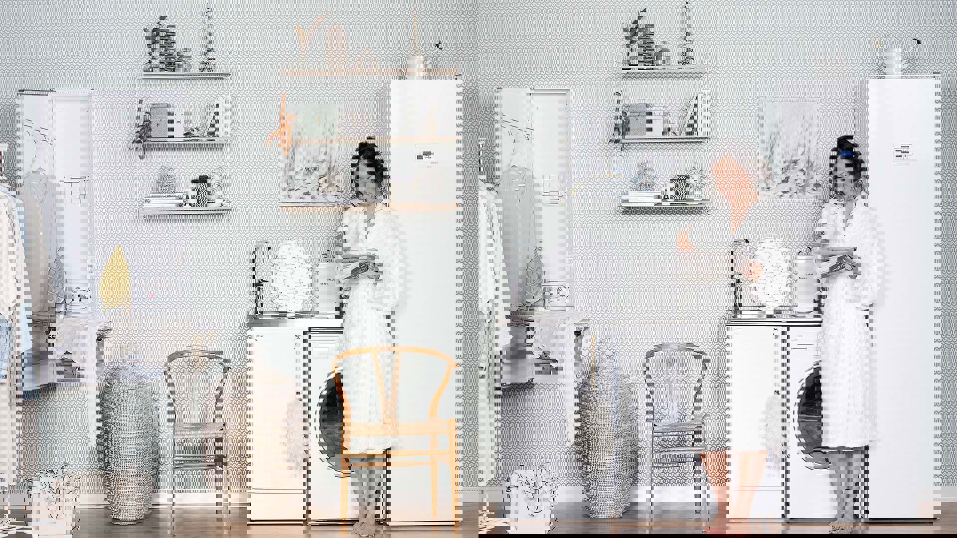 En kvinna i vit frottérock med en kaffekopp och en tidning står framför ett vitt torkskåp från Nimoverken i en tvättstuga. 